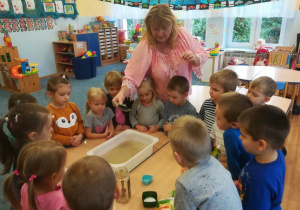 Dzieci stoją dookoła stołu nauczycielka wsypuje pieprz do miski z wodą
